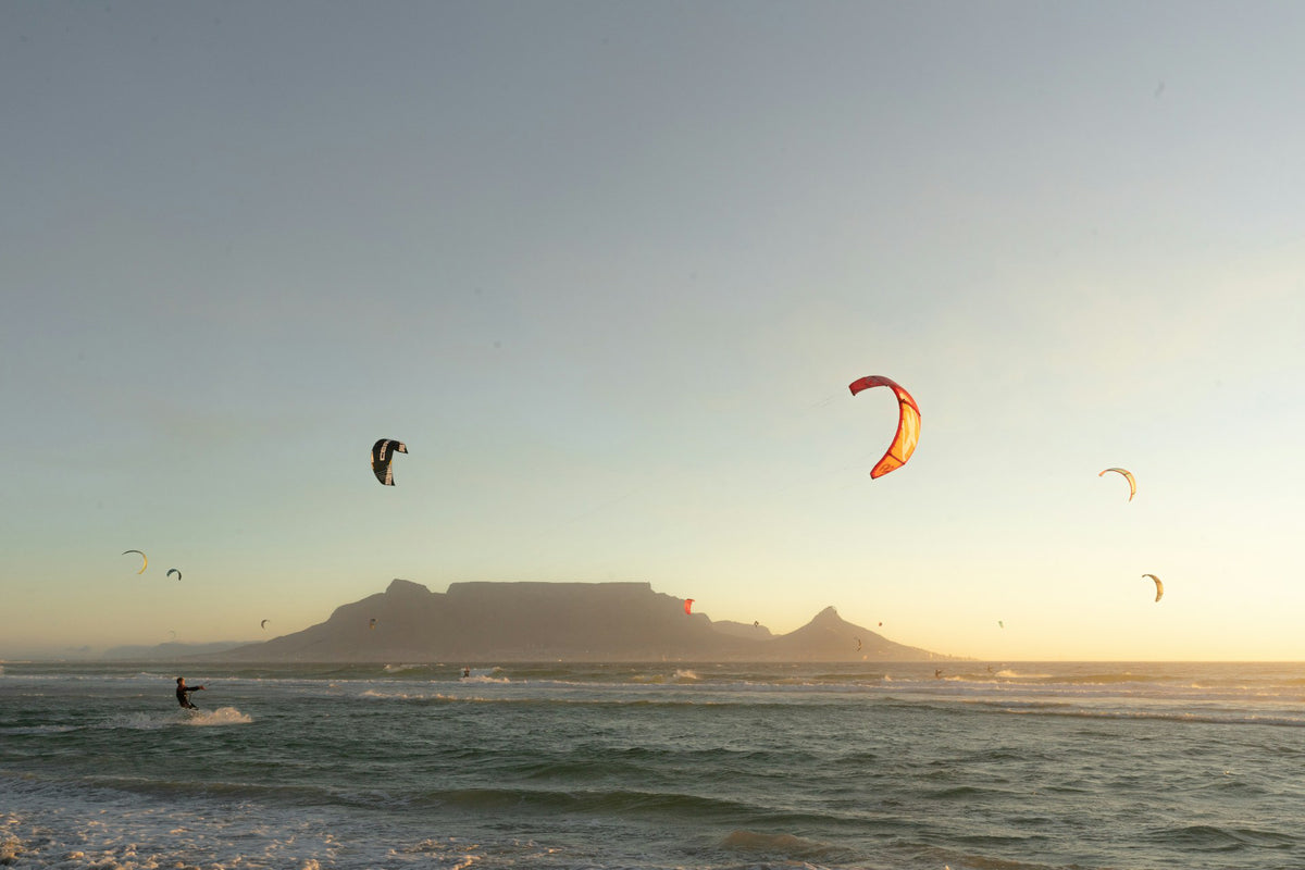 A wide outdoor seascape at sunset with several kitesurfers on the water in the foreground and a large mountain or mesa silhouette in the background. The sky is pale blue to warm beige near the horizon, and the water is dark blue-gray.