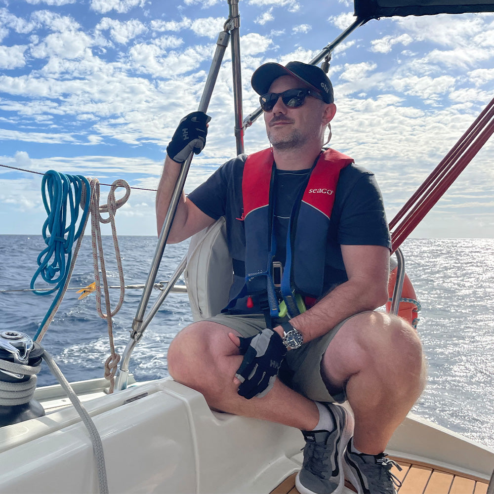 Man on a sailboat wearing a life jacket and sunglasses with a blue sky and ocean background.