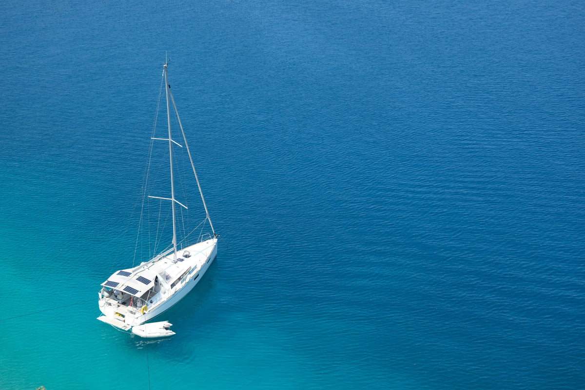 A white sailboat with its sails down floats on clear turquoise blue water, photographed from a high-angle aerial perspective.